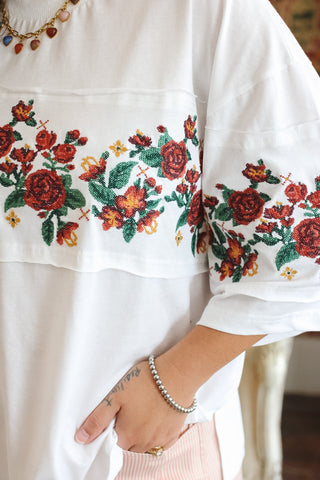 A close-up of a white embroidered top with red and yellow floral patterns and green leaves across the chest and sleeve, worn by a model with a beaded bracelet and hand in pocket.
