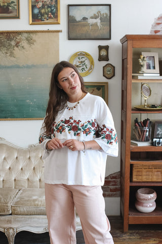 A woman modeling a white tee with red and green floral embroidery on the chest and sleeves, paired with light pink striped pants, standing in a vintage-style room with a tufted sofa and wooden cabinet.