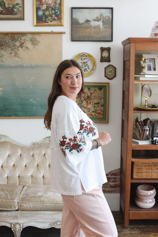 A woman wearing a white long-sleeve top with red and green floral embroidery on the sleeves, paired with light pink striped pants, standing in a decorated room with vintage furniture and framed art.