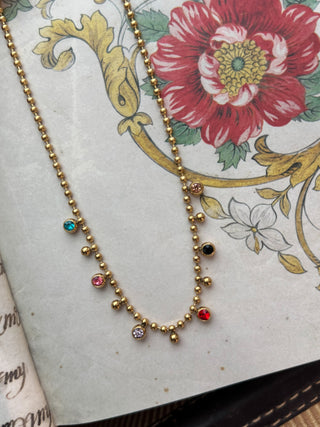 A gold beaded necklace with small colorful gemstone accents, displayed on an open vintage book with a floral illustration.
