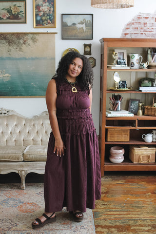 Woman wearing a sleeveless maroon maxi dress with a smocked bodice, standing in a vintage-decorated room.