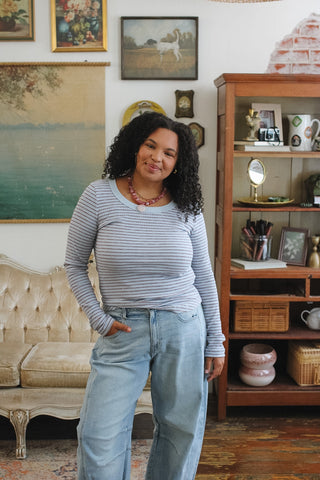 A woman with curly black hair stands in a cozy, vintage-style room wearing a light blue and brown striped long-sleeve top and light-wash blue jeans.