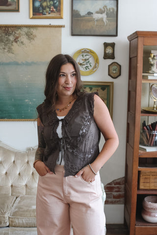A woman stands in a decorated room wearing a sleeveless dark brown lace ruffled top over a white undershirt and light pink pants.