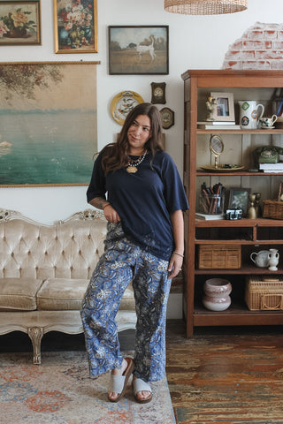 A woman stands in a styled room wearing a navy blue t-shirt and blue floral wide-leg pants, paired with white slide sandals. The background features a vintage tufted sofa, a wooden bookshelf, and framed wall art.