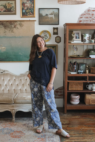 A woman stands in a vintage-style room wearing a navy blue top and blue floral wide-leg pants, paired with beige slide sandals. The room features a tufted cream sofa, a wooden bookshelf, and framed wall art.