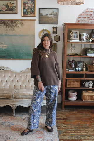 A woman stands in a vintage-style room wearing a brown short-sleeve poncho top and blue floral wide-leg pants. The room features a tufted cream sofa, a wooden bookshelf, and framed artwork on the walls.