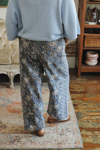 A person viewed from behind wearing wide-leg blue patterned trousers with a light blue knit top, standing on a patterned rug in an indoor room with wooden furniture.