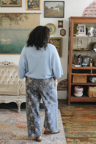 A person viewed from behind wearing a light blue ribbed knit top and blue floral wide-leg pants, standing in a vintage-style room with a wooden cabinet, tufted sofa, and framed wall art.