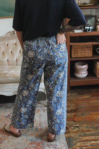 A person wearing loose, wide-leg pants with an intricate blue, gray, and beige paisley pattern, paired with a black top and brown sandals, standing in a vintage-style room.