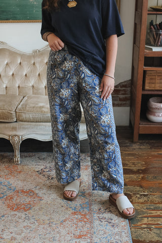 A person wearing loose blue paisley print pants with a navy t-shirt and white slide sandals, standing in a vintage room with a tufted sofa and wooden shelving.