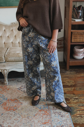 A person wearing loose brown knit top and blue paisley print wide-leg pants with dark flats, standing in a vintage-style room with a tufted sofa and wooden shelving.