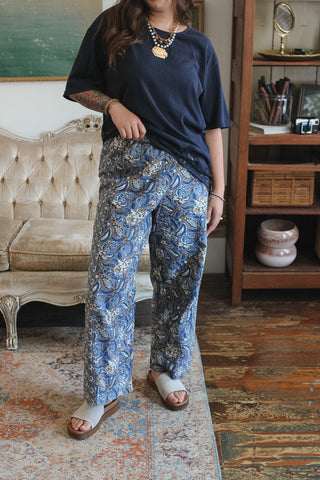 A person standing indoors wearing loose dark blue short-sleeve top and wide-leg blue patterned pants with white slide sandals, photographed in a styled room with a tufted sofa and wooden shelving.
