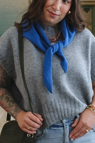 A woman stands wearing a gray knit sleeveless sweater vest, light blue jeans, and a bright blue wool knit triangle neck scarf.