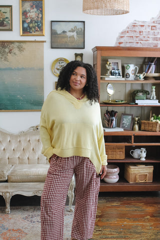 A woman wearing a pale yellow oversized knit hooded sweater and red-and-white gingham pants stands in a decorated room with paintings, a tufted bench, and a wooden bookshelf.