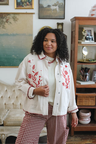 A woman wearing a cream-colored embroidered jacket with red floral patterns, a cream top, and red-and-white gingham pants, standing in a vintage-style room with a wooden cabinet and framed art in the background.
