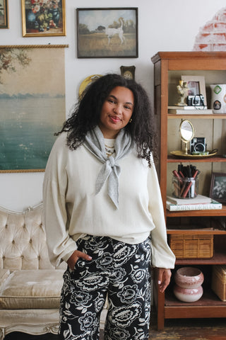 Woman wearing a cream sweater with a gray scarf and black floral-print pants standing in a vintage-style room.