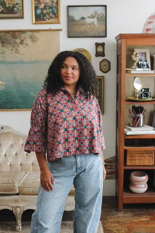 A woman wearing a colorful floral blouse and light blue jeans stands in a vintage-decorated room.