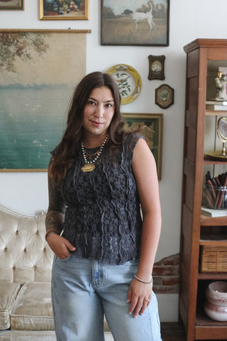 A woman wearing a dark gray sleeveless lace top and light blue jeans, standing in a decorated room with framed art, a tufted sofa, and a wooden cabinet.