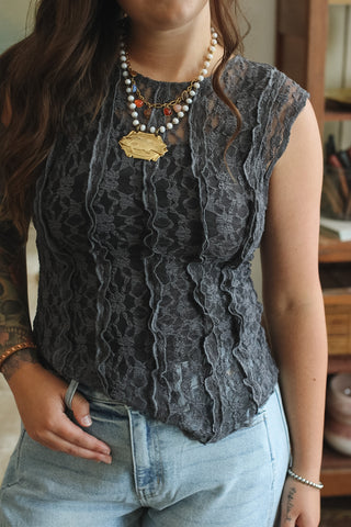 A woman wearing a sleeveless dark gray lace top with vertical ruffles, paired with light blue jeans and layered necklaces, shown in a warm indoor setting.