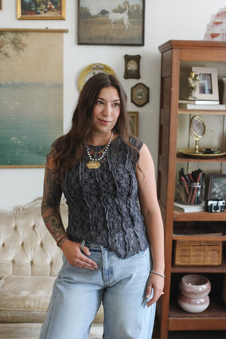 A woman with long brown hair stands in a vintage-style room wearing a dark gray lace sleeveless top and light blue jeans.