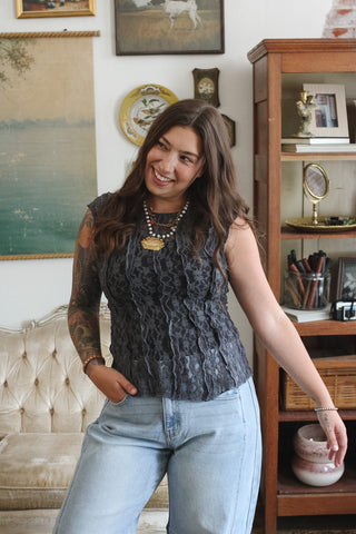 A woman wearing a dark gray lace sleeveless top and light blue jeans, standing in a decorated room with a wooden cabinet and framed art in the background.