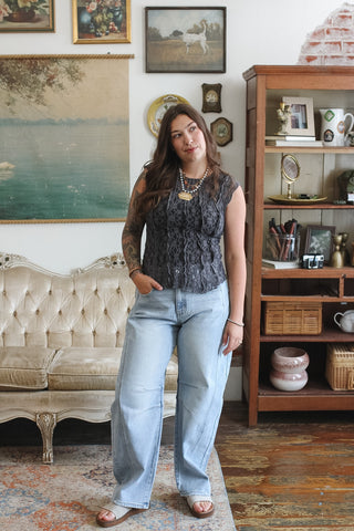 A woman stands in a vintage-style room wearing a sleeveless black lace-patterned top and light-wash wide-leg jeans.