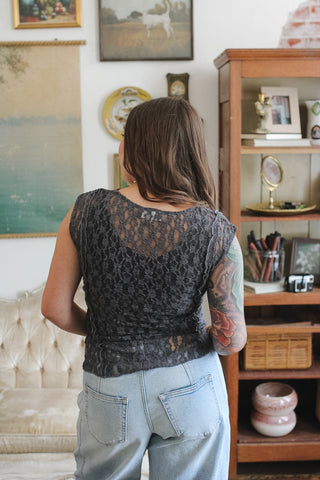 A woman viewed from behind wearing a sleeveless black lace top and light blue jeans in a decorated room with framed art and a wooden bookshelf.