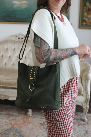 A woman wearing a white knit top and red-and-white gingham pants carries an olive green suede shoulder bag with a studded strap detail and tassel, standing in a decorated room with a tufted sofa in the background.