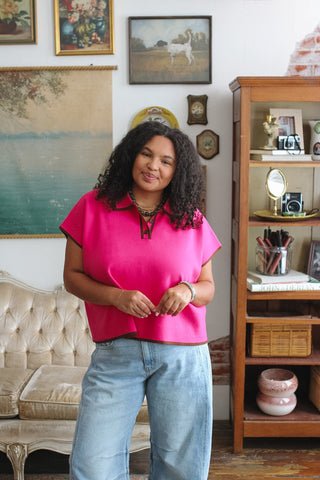 Woman wearing a bright pink short-sleeve top and light blue jeans standing in a vintage-style room.