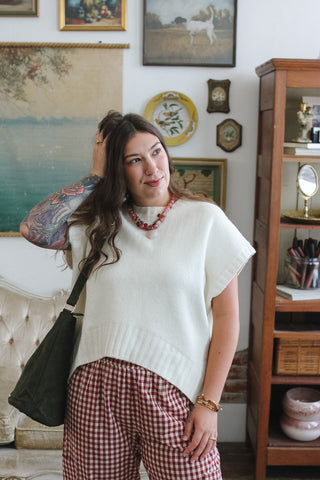 A woman posing in a cozy room wearing a cream knit cap sleeve sweater and red-and-white gingham pants, with a dark green shoulder bag.