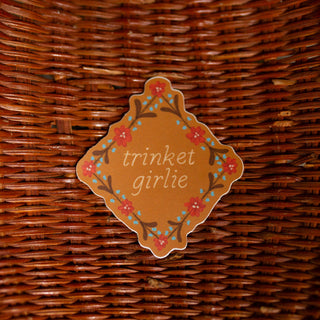 A sticker with the words 'trinket girlie' in white script, surrounded by a floral border in red, and blue on a warm brown background.