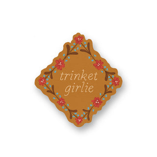 A sticker with the words 'trinket girlie' in white script, surrounded by a floral border in red, and blue on a warm brown background.