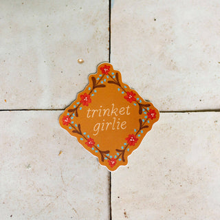 A sticker with the words 'trinket girlie' in white script, surrounded by a floral border in red, and blue on a warm brown background.