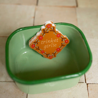 A sticker with the words 'trinket girlie' in white script, surrounded by a floral border in red, and blue on a warm brown background.
